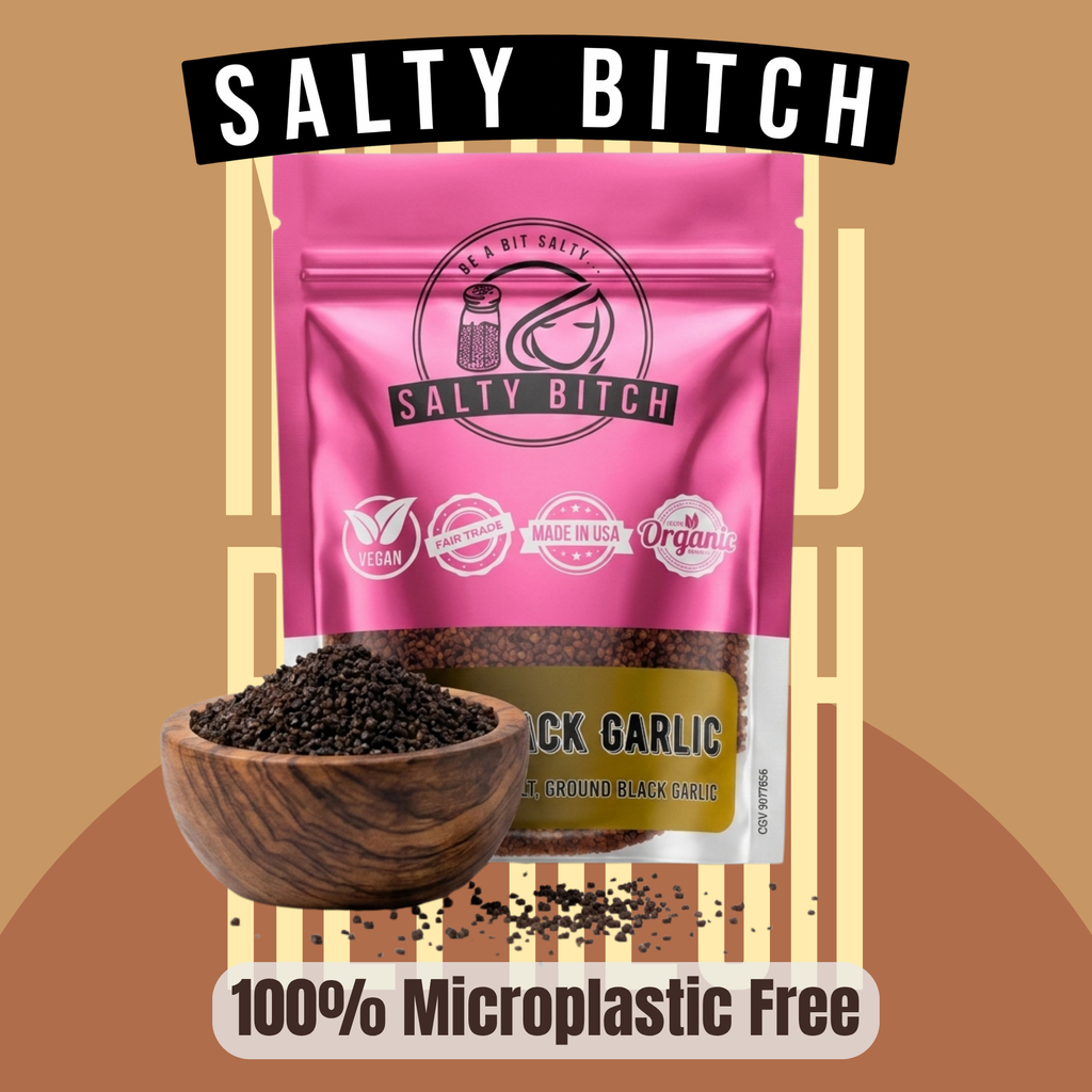 A pink stand-up pouch of Salty Bitch Black Garlic with a wooden bowl of dark ground garlic in front, on a tan graphic background with the words 'SALTY BITCH' at the top and '100% Microplastic Free' at the bottom.