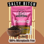 A pink stand-up pouch of Salty Bitch Black Garlic with a wooden bowl of dark ground garlic in front, on a tan graphic background with the words 'SALTY BITCH' at the top and '100% Microplastic Free' at the bottom.