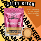 A pink stand-up pouch of Salty Bitch Habanero Sea Salt with a clear window showing the ground habanero salt inside, alongside a wooden bowl filled with the salt. The background is a bold graphic with orange and yellow diagonal blocks and large black text repeating 'HABANERO'.